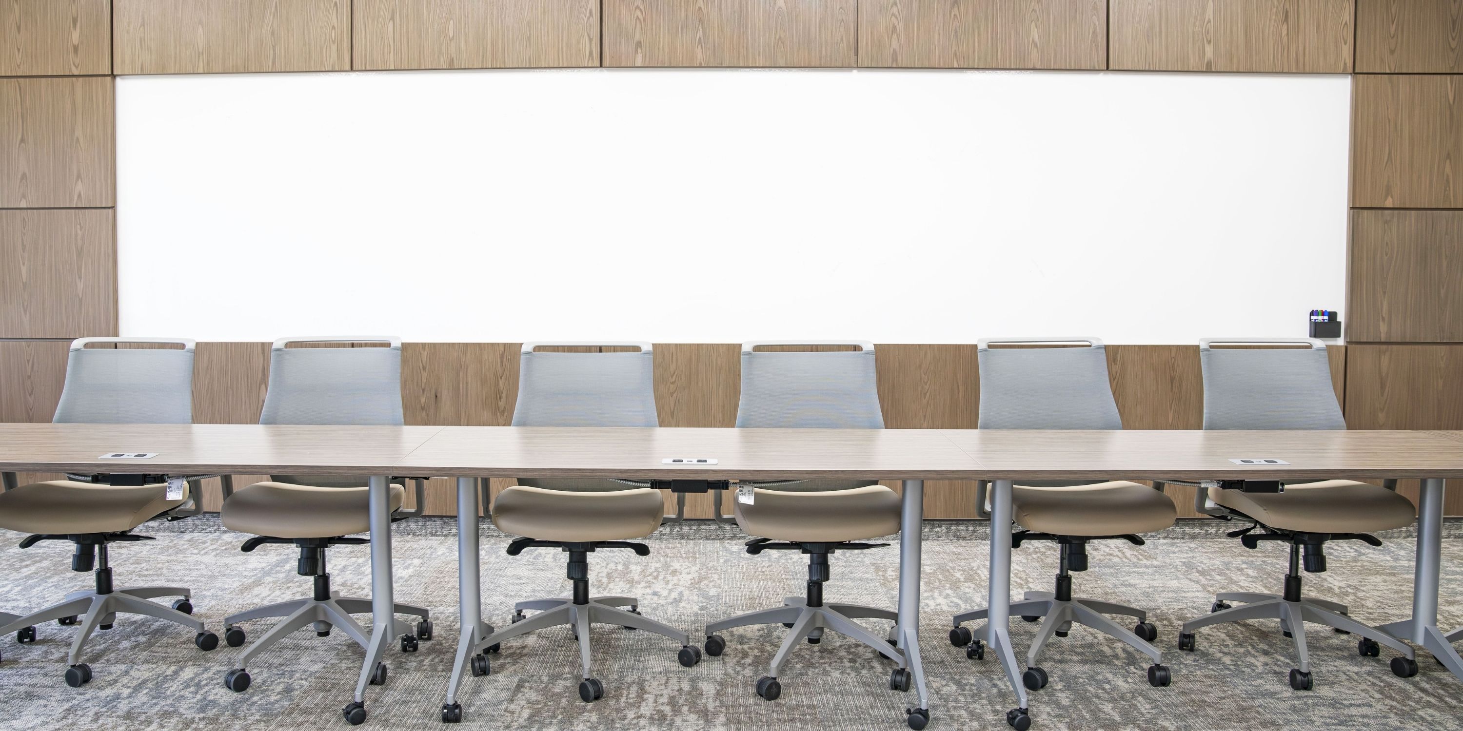 A modern conference room with a long wooden table and eight beige office chairs on gray carpet. A large whiteboard on a wooden wall backdrop conveys professionalism.