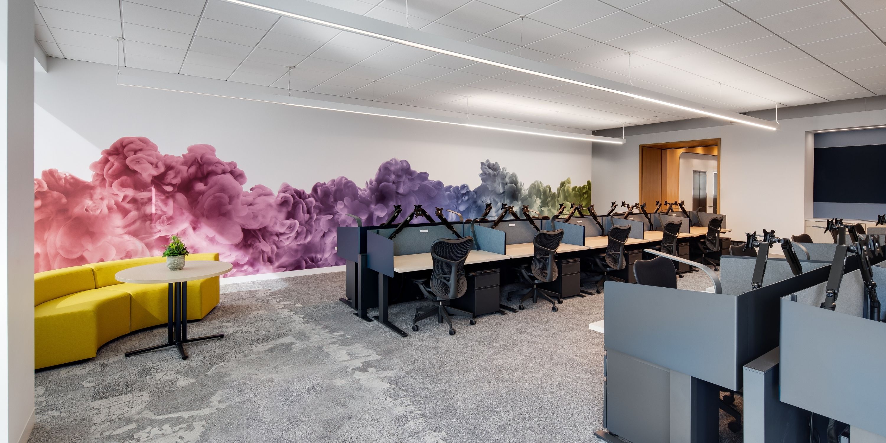 A modern office space features a vibrant mural of colorful ink clouds on the wall, with rows of neat desks and a cozy yellow sofa creating a lively atmosphere.