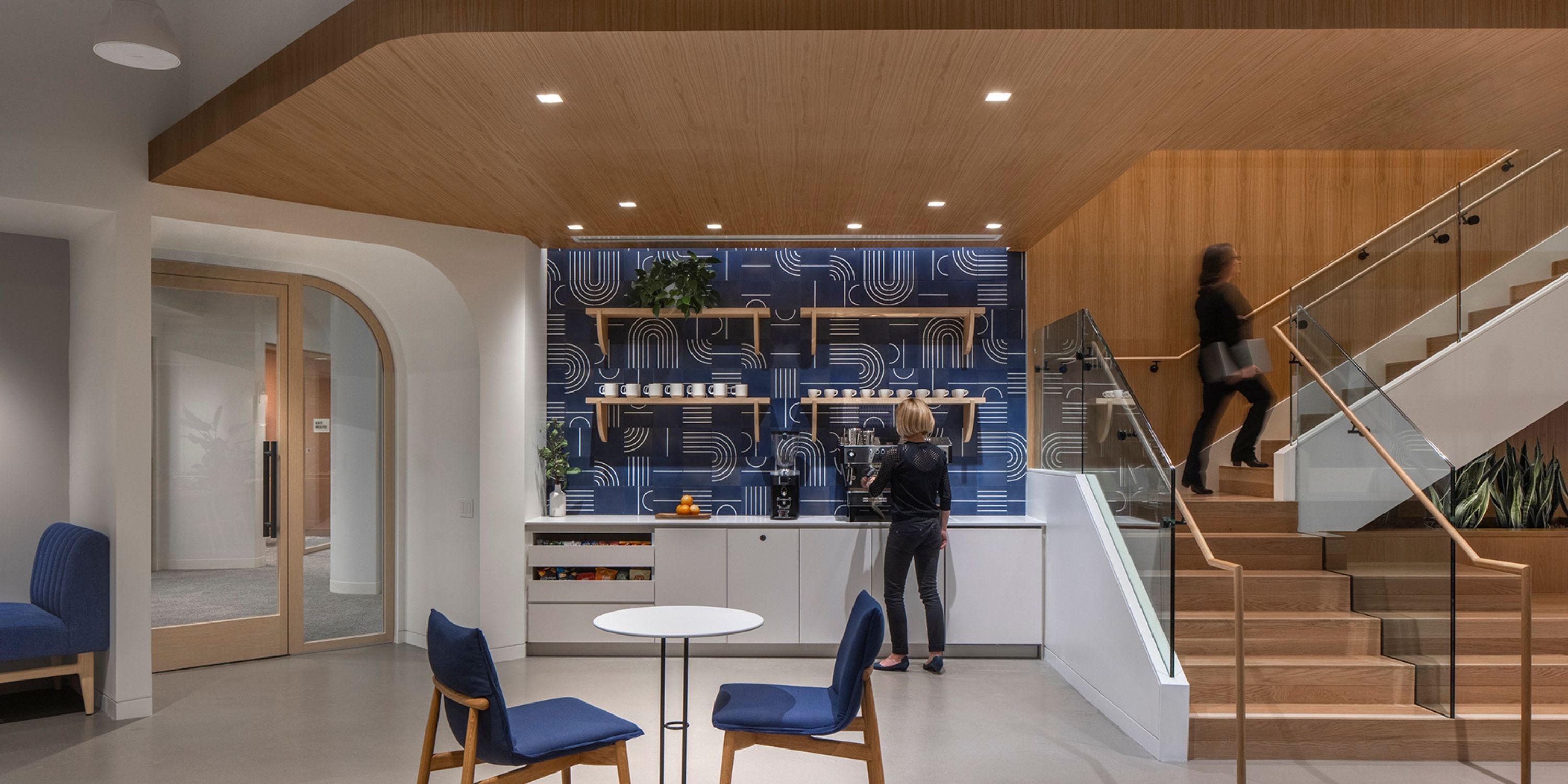 Modern office lounge with a coffee station, blue patterned wall, and plants. Two blue chairs by a round table; a woman walks up wooden stairs.