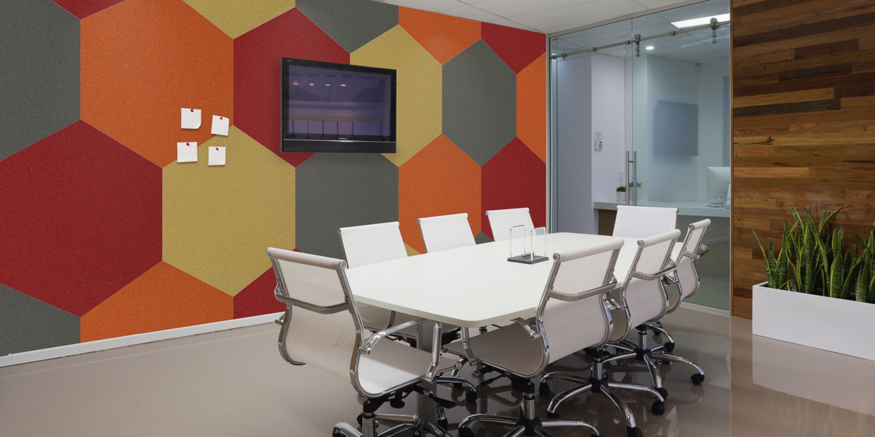 A modern conference room with a long white table, white chairs, and colorful hexagonal wall patterns. A TV is mounted on the wall. A plant adds greenery.