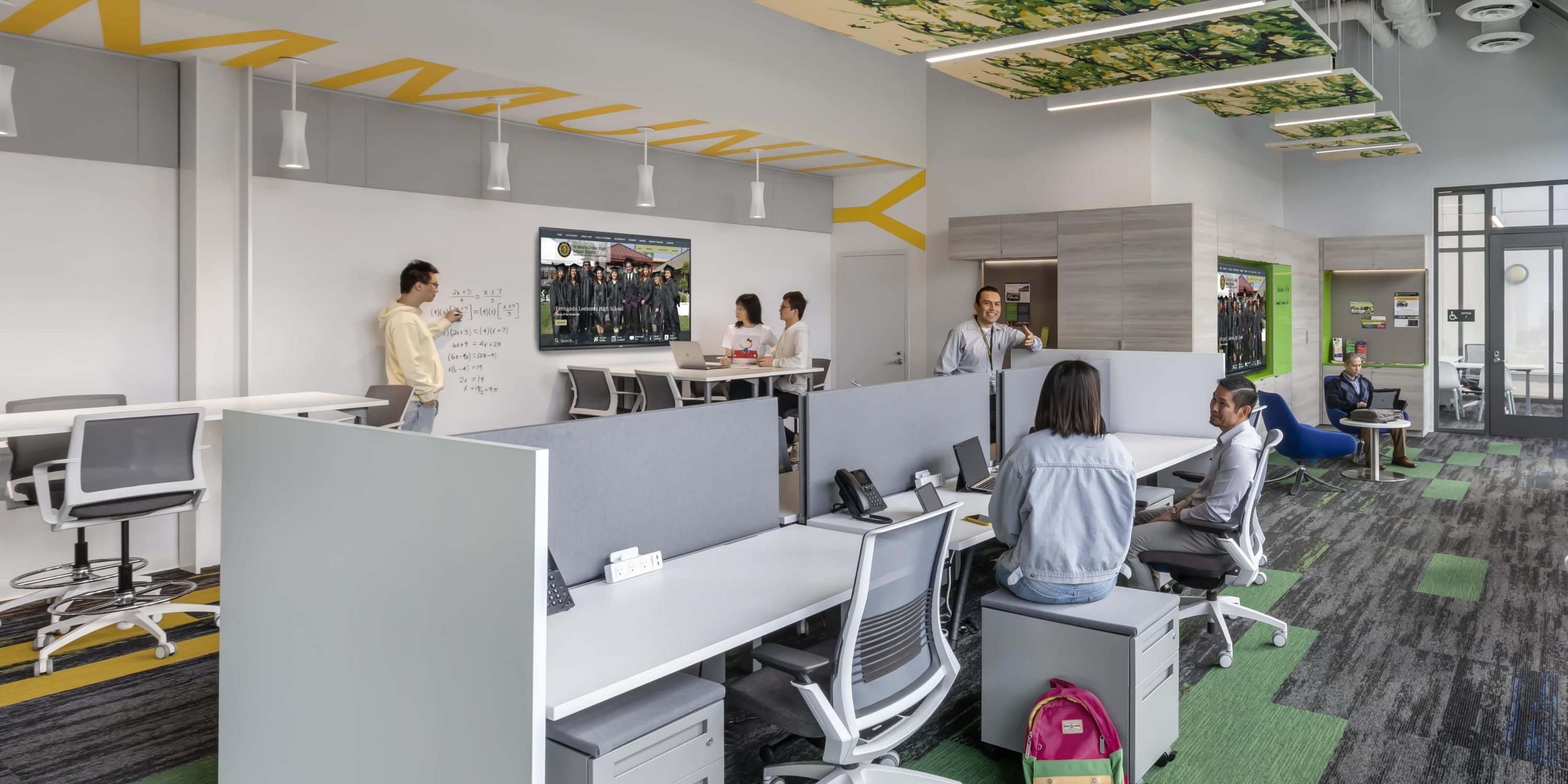 Modern office space with people collaborating. Open-plan design features desks, a whiteboard, and a digital screen, conveying a productive atmosphere.