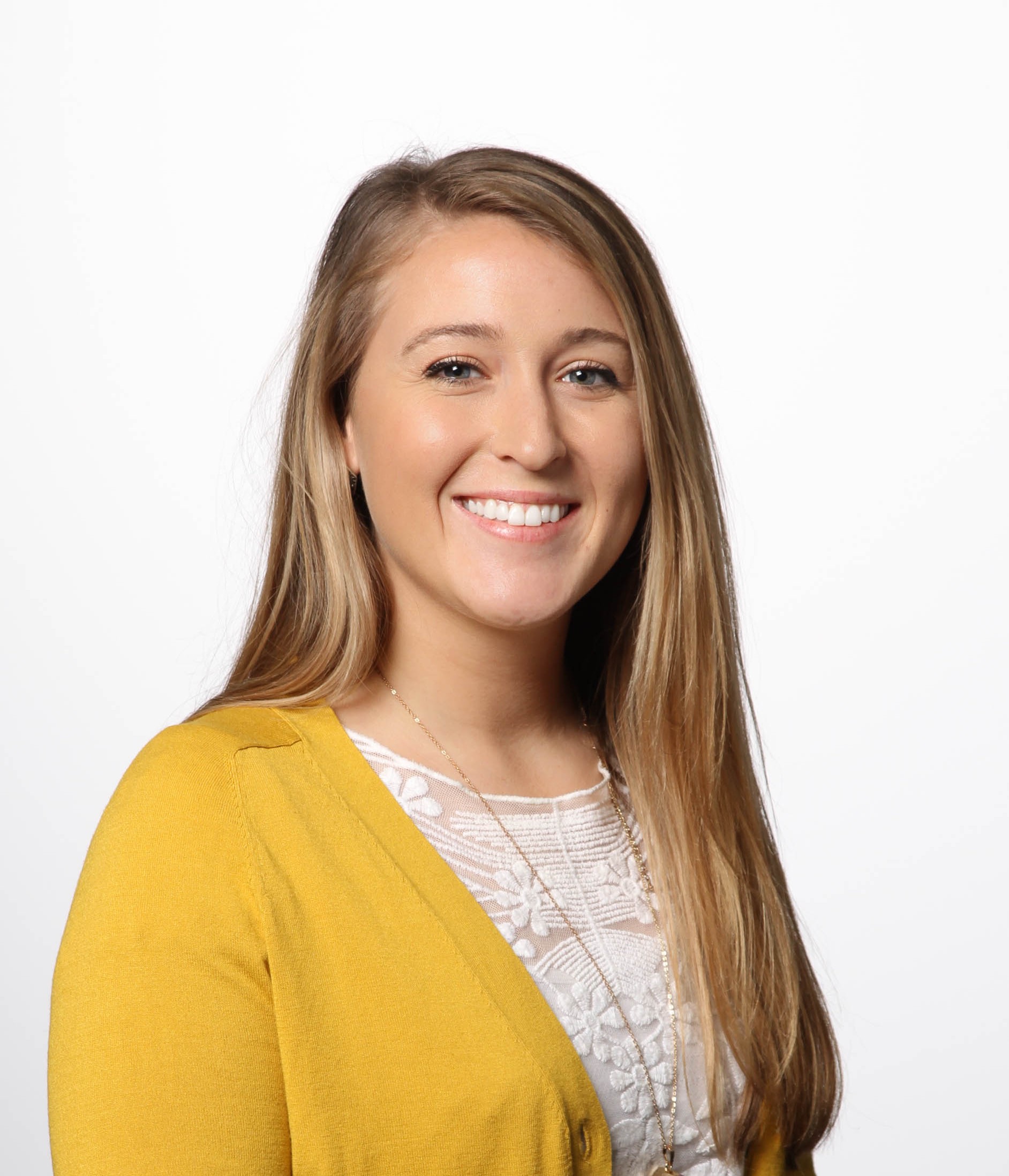 Smiling woman with long straight blonde hair, wearing a mustard yellow cardigan over a white lace blouse and a pendant necklace, posed in front of a plain white background.