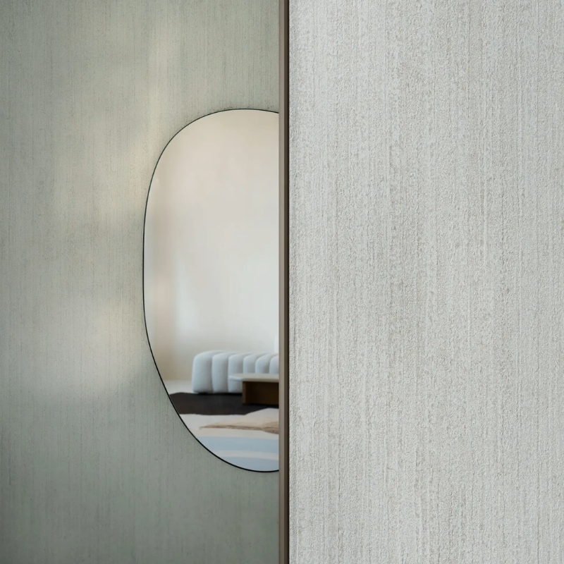A minimalist room with light, textured walls features an elongated oval mirror. The mirror reflects a modern white couch, conveying a calm, contemporary vibe.