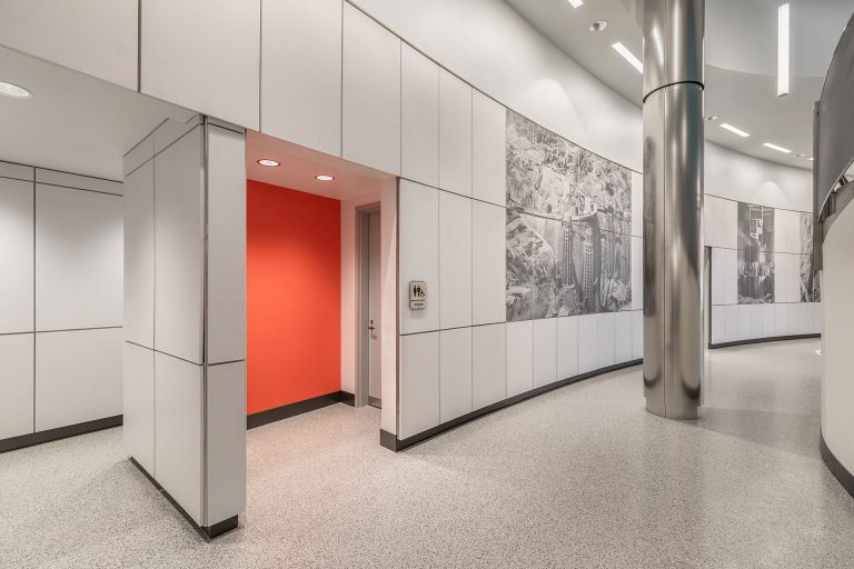 Curved corridor with white paneled walls and photo murals, featuring a vibrant red restroom entrance. Sleek metallic column enhances modern aesthetic.