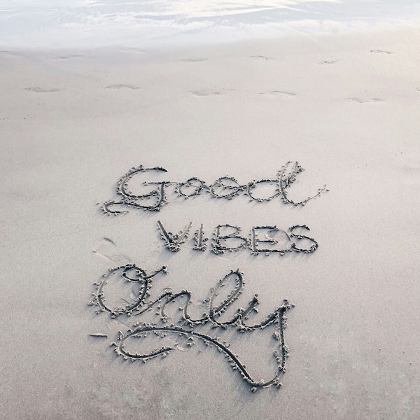 "Good Vibes Only" is written in cursive in beach sand, near gentle waves and footprints, conveying a peaceful, positive, and relaxed atmosphere.