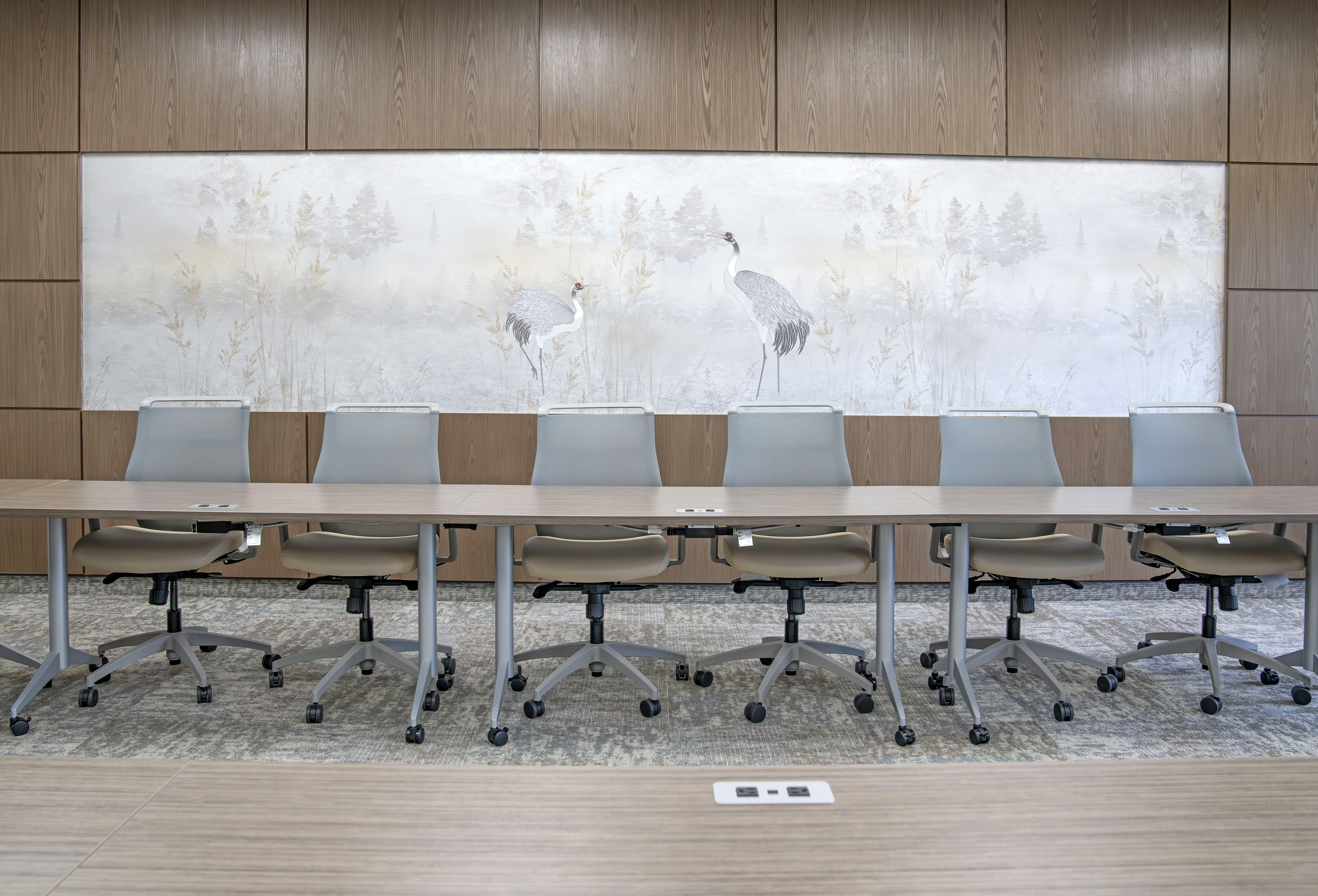 A modern conference room with eight gray office chairs around a long wooden table. Behind, a wall mural features two cranes in a serene landscape.