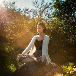 A woman meditates in lotus position on a rock, surrounded by lush greenery. Sunlight filters through trees, creating a peaceful, serene atmosphere.