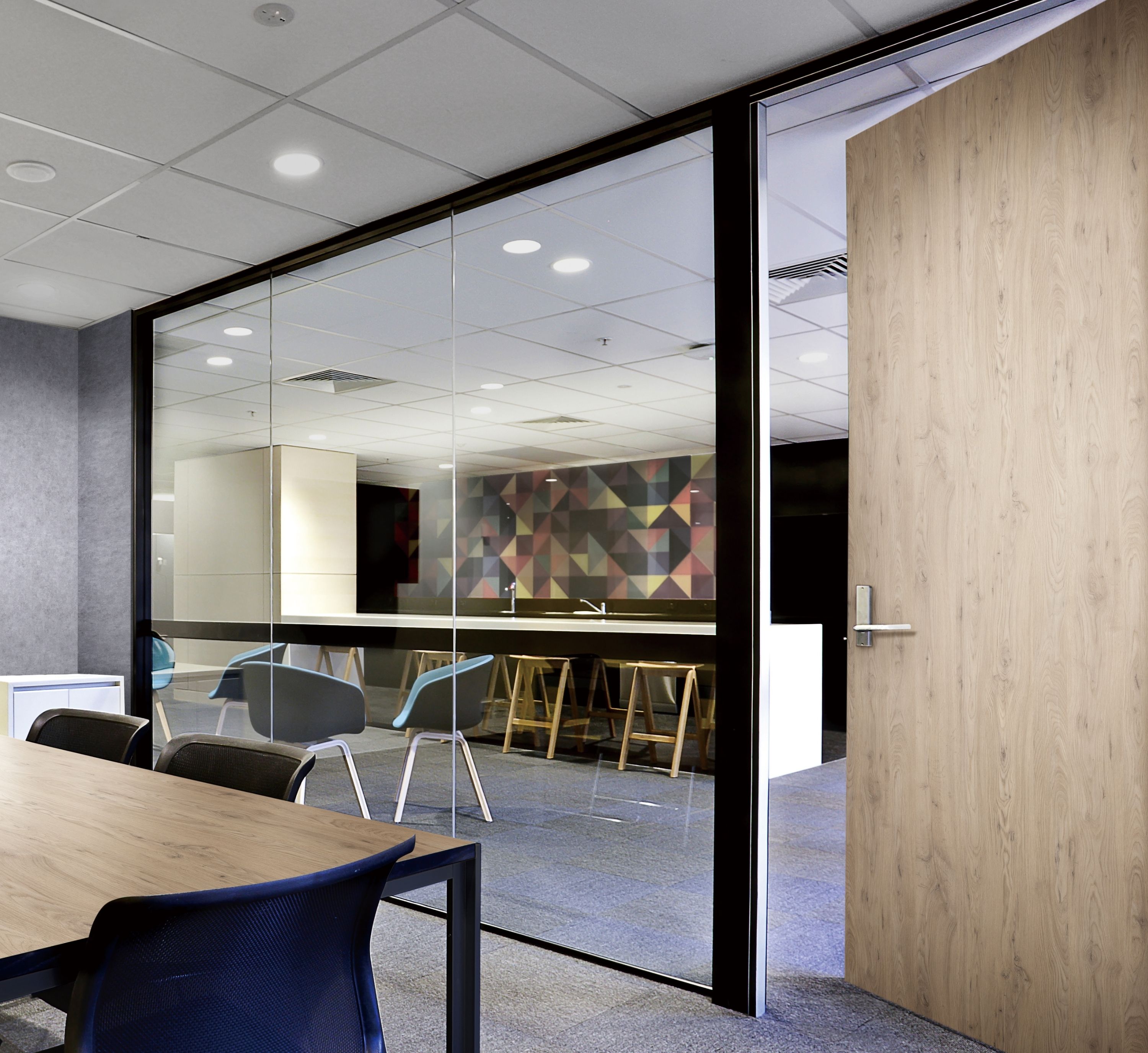 A modern conference room with a wooden table and black chairs. A glass wall reveals a colorful, geometric-patterned lounge with blue chairs and bar stools.