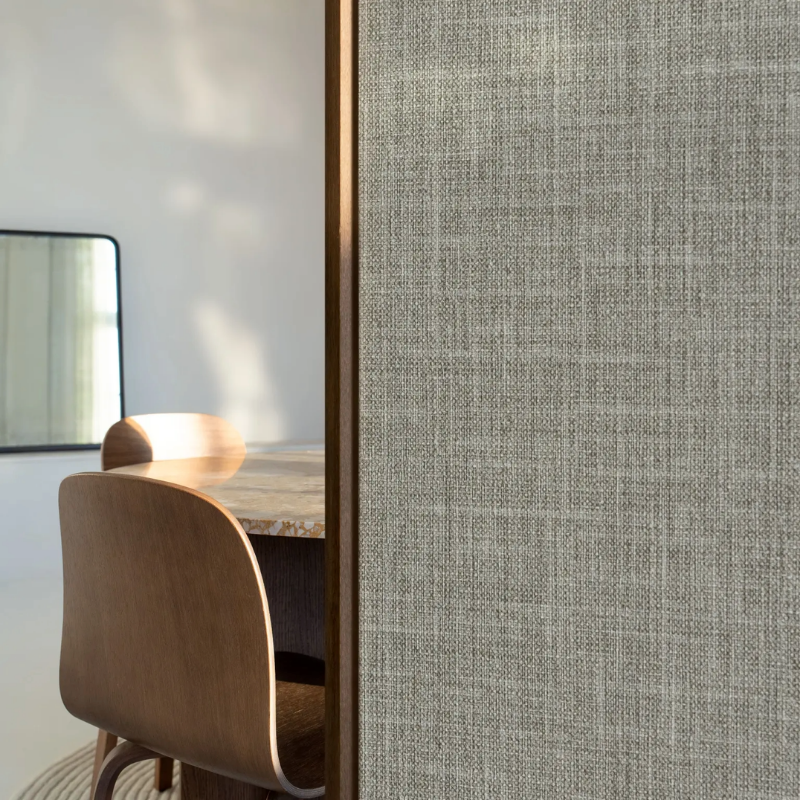 Wooden chair and table partially visible behind a textured gray partition in a softly lit room; a mirror reflects light, creating a calm, modern vibe.