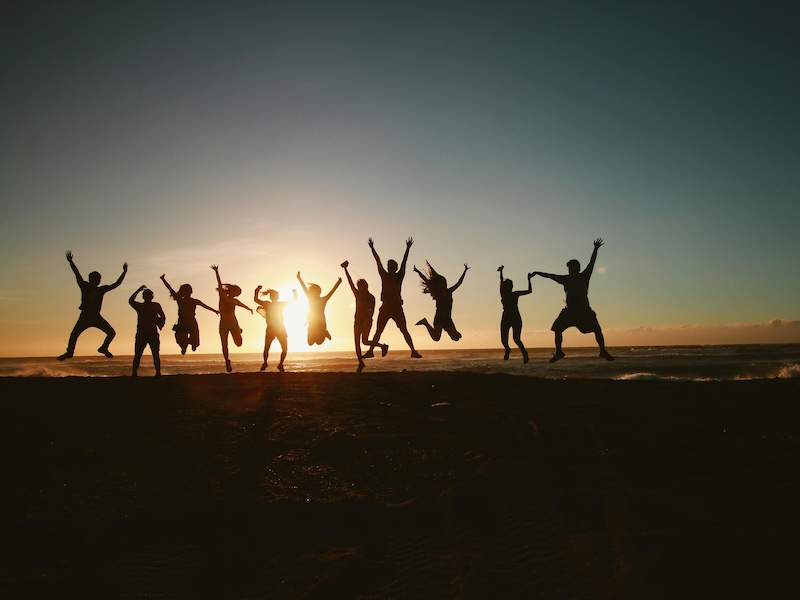 Silhouetted group of people jumping joyfully on a beach at sunset. The sun is low on the horizon, casting a warm glow and vibrant sky.