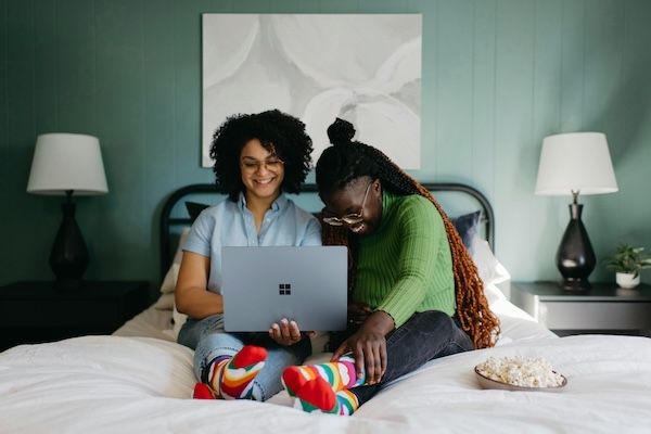 Two women sit on a bed together, smiling and looking at a laptop. Both are wearing colorful rainbow-striped socks, and there is a bowl of popcorn on the bed beside them. A modern bedroom setup with dark lamps and a large abstract painting is in the background.