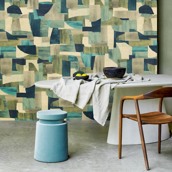 Modern dining setting with abstract geometric wallpaper in earthy tones. Features a sleek white table, wooden chair, blue stool, and a gray cloth.