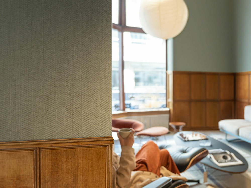 HERITAGE-WOOD-II-(2).png Cozy living room with wood paneling; someone in a beige sweater holds a mug, seated on a modern chair. Soft light filters through large w.indows, creating a warm ambiance