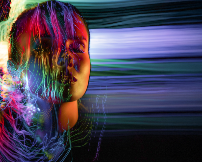 A woman's face is illuminated with vibrant, multicolored light streaks, creating a dreamlike, ethereal effect against a blurred, dark background.