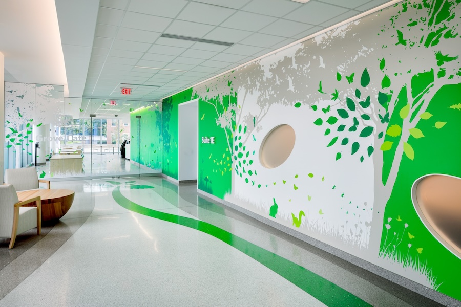Bright hallway with a nature-themed mural featuring green trees, leaves, and animals. Glass doors lead to a seating area, creating a serene atmosphere.