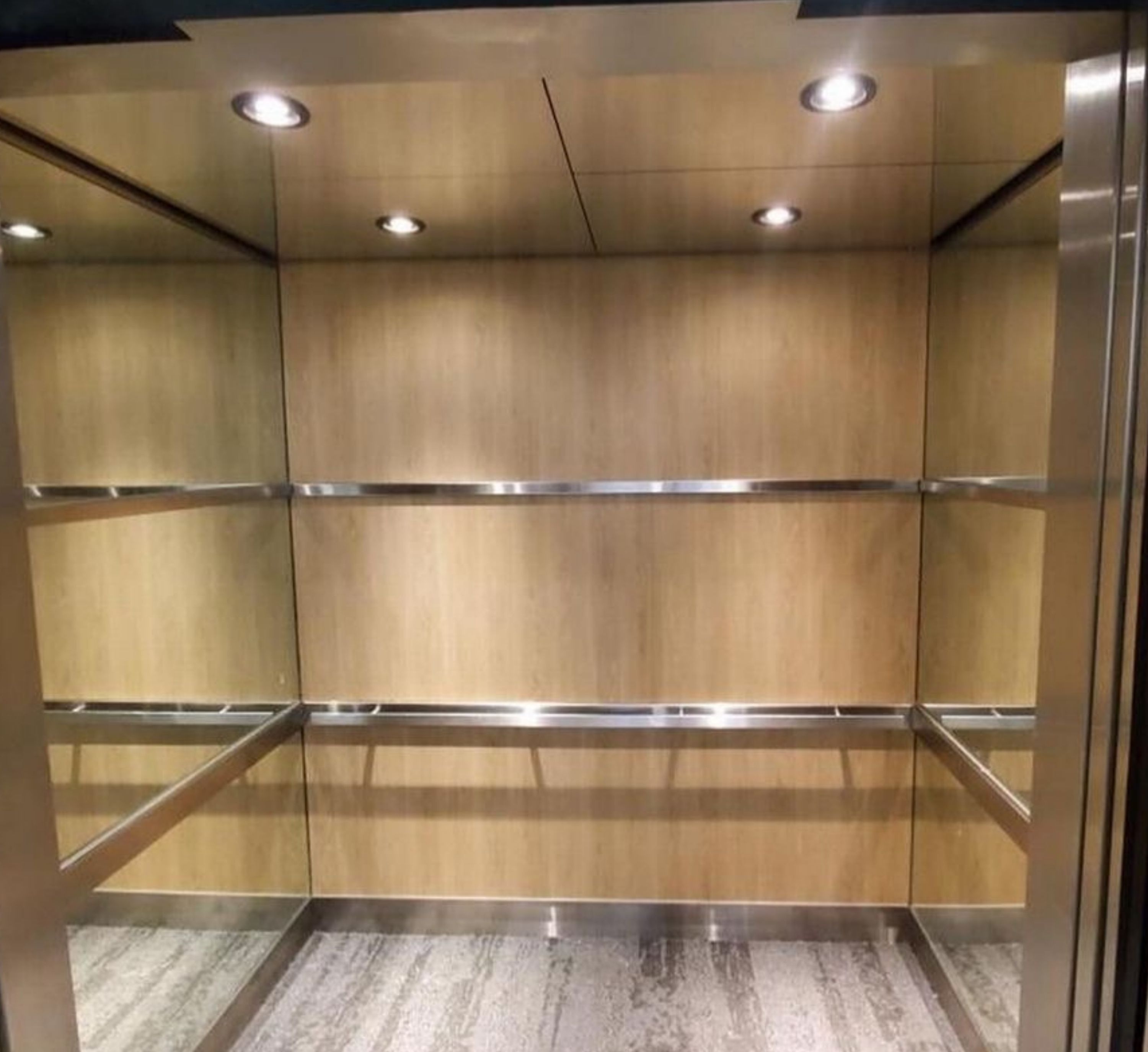 Empty elevator interior with wood panel walls, metallic accents, and recessed lighting. Gray textured floor adds modern touch. Calm and quiet tone.